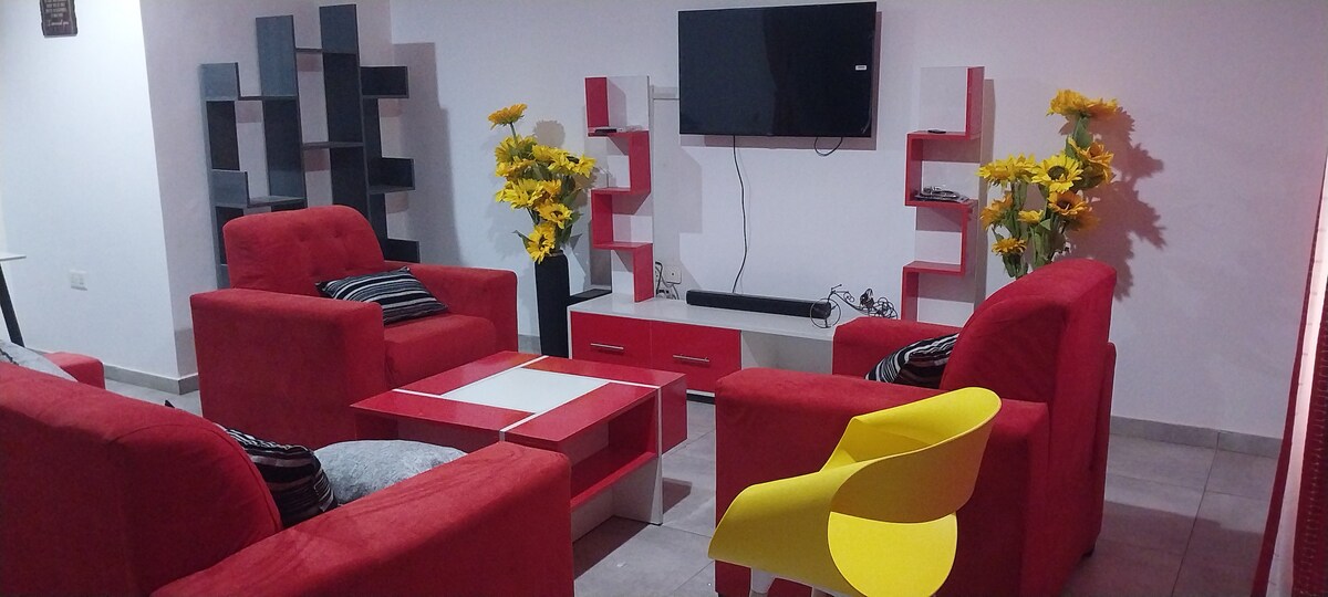 A seating area is arranged with vibrant red sofas surrounding a coffee table. A white entertainment unit is positioned below a mounted television. Yellow flowers are placed in decorative vases, adding a touch of color. Gray shelves complement the modern aesthetic.