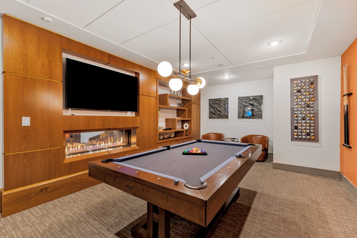 A game room is presented, featuring a billiard table in the center surrounded by comfortable seating. A contemporary electric fireplace is integrated into the wooden cabinetry, while a large television is mounted on the wall. Unique wall art adds character to the room.