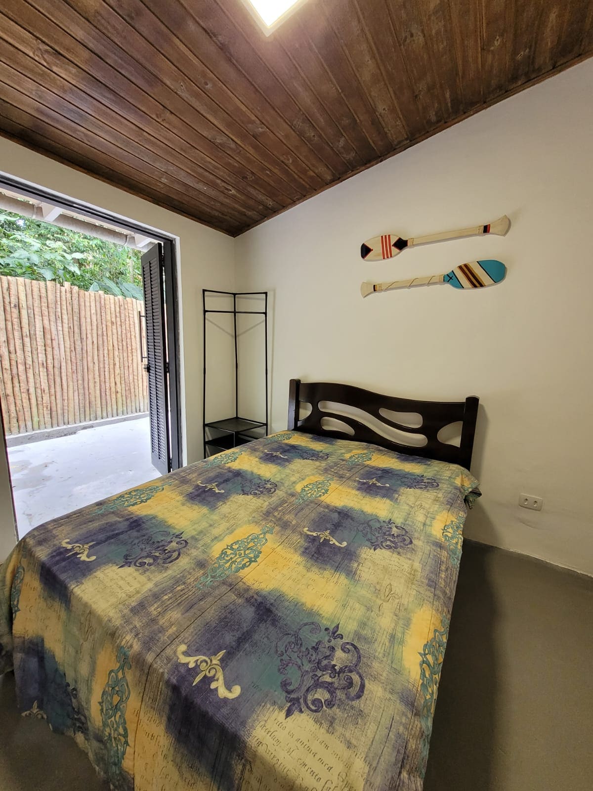 A cozy bedroom features a spacious bed adorned with a colorful patterned blanket. Two decorative wooden paddles are mounted on the wall above the bed. Light enters through an open door, leading to a private outdoor space, creating a bright and airy atmosphere.
