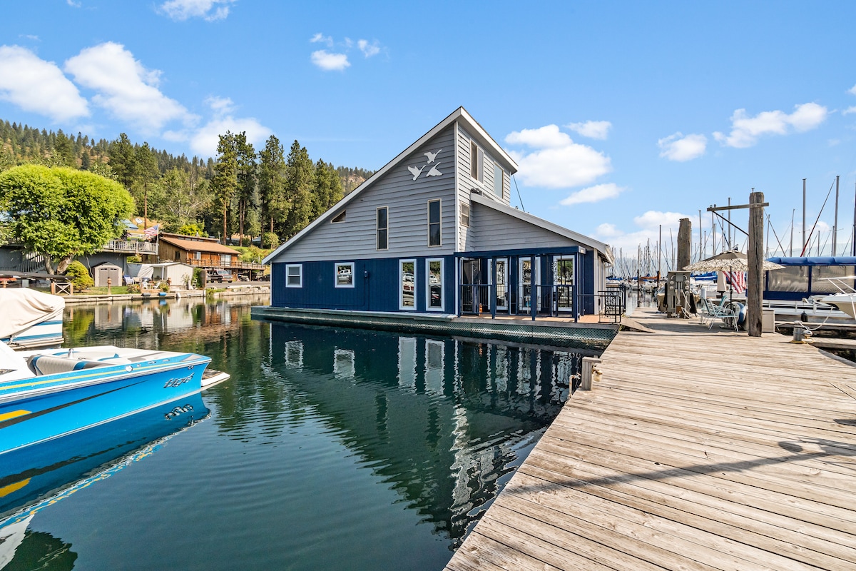 A modern floating home is showcased, featuring a unique A-frame design with blue siding. Surrounded by water, the property has large windows reflecting the serene lake, with a wooden dock extending from the shore, providing access to parked boats and scenic views.