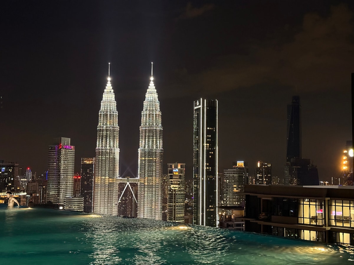 A panoramic view of the iconic Petronas Towers is captured at night, illuminated against the skyline. The scene features an infinity pool in the foreground, extending towards the towers, creating a striking reflection of the vibrant city lights.