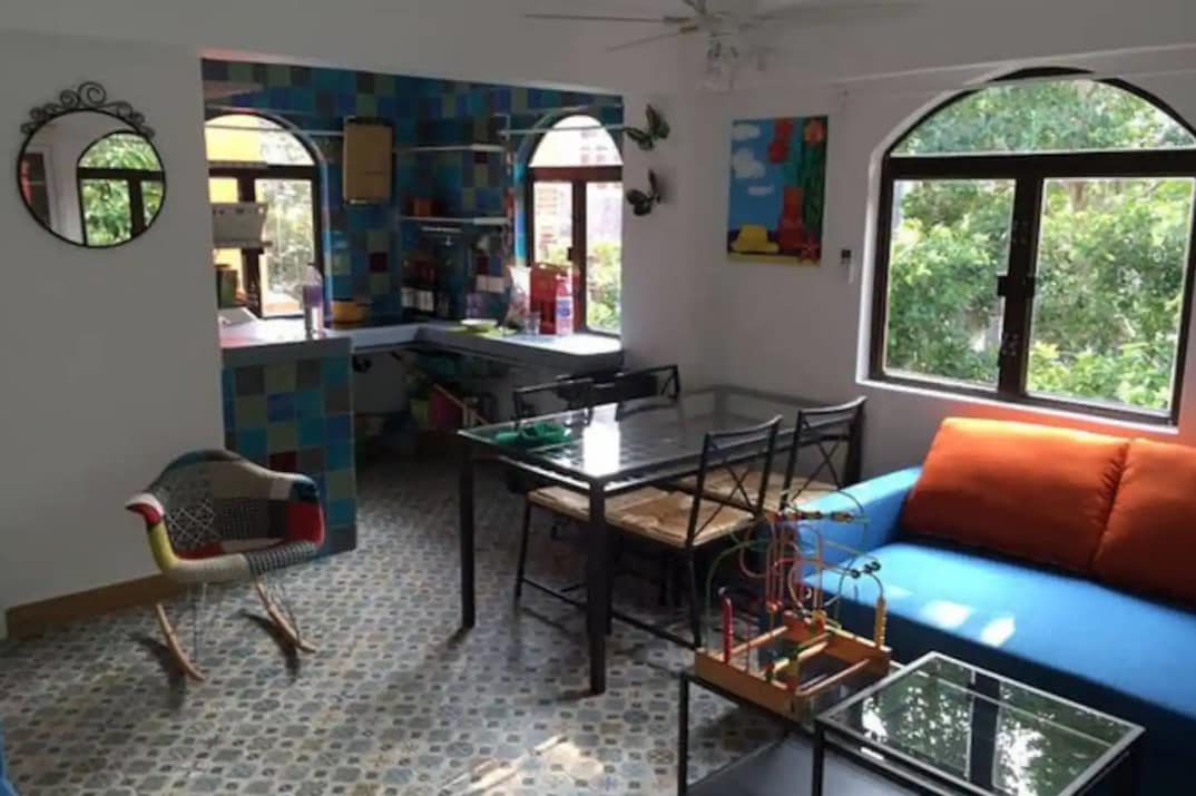 A bright and airy living space features colorful tile accents and large windows that invite natural light. A blue sofa and patterned chair are visible, along with a glass dining table. A kitchen area with shelves is seen in the background, surrounded by greenery.