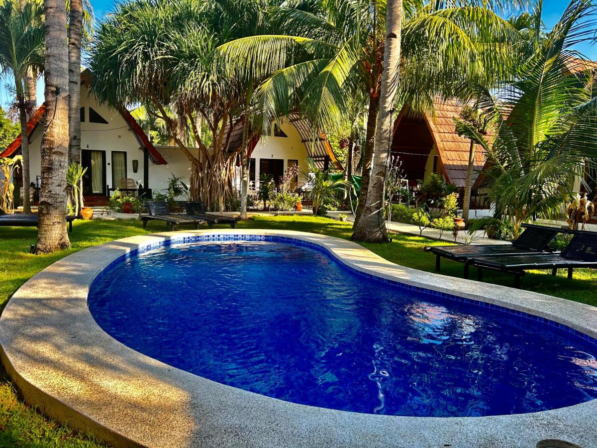 A tranquil swimming pool is surrounded by palm trees and tropical foliage, inviting relaxation. The outdoor area features lounge chairs available for sunbathing, while charming bungalow-style buildings are visible in the background, creating a serene and lush environment.