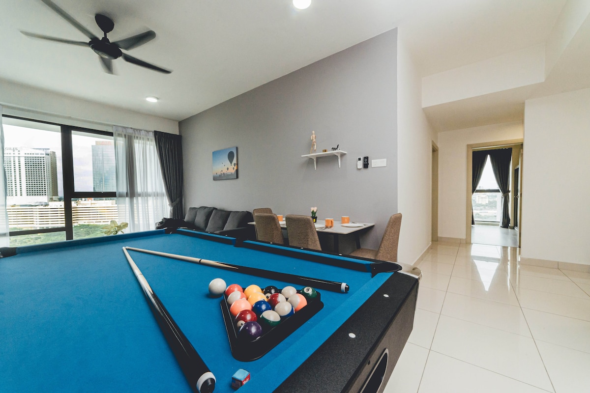 A spacious living area features a pool table with a set of colorful balls arranged on it. A comfortable sofa is positioned nearby, along with a dining table set for four. Large windows allow natural light to illuminate the space.