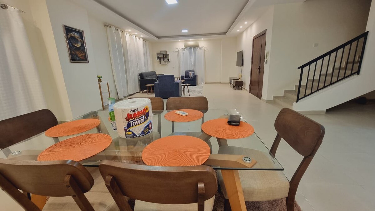 A dining area is equipped with a glass table surrounded by four wooden chairs. Bright orange placemats adorn the table. In the background, a seating area can be seen, featuring a sofa and a television. Soft, natural light enters through sheer curtains, enhancing the overall openness.