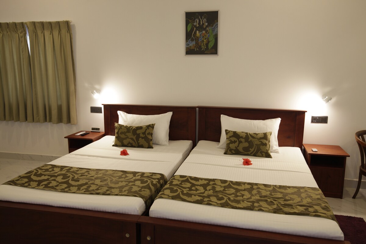 A comfortable sleeping area features two spacious beds, each adorned with decorative pillows. A small flower is placed on the bedding. Soft lighting is provided by wall sconces, and a piece of artwork is displayed above the beds. Light curtains frame a window.