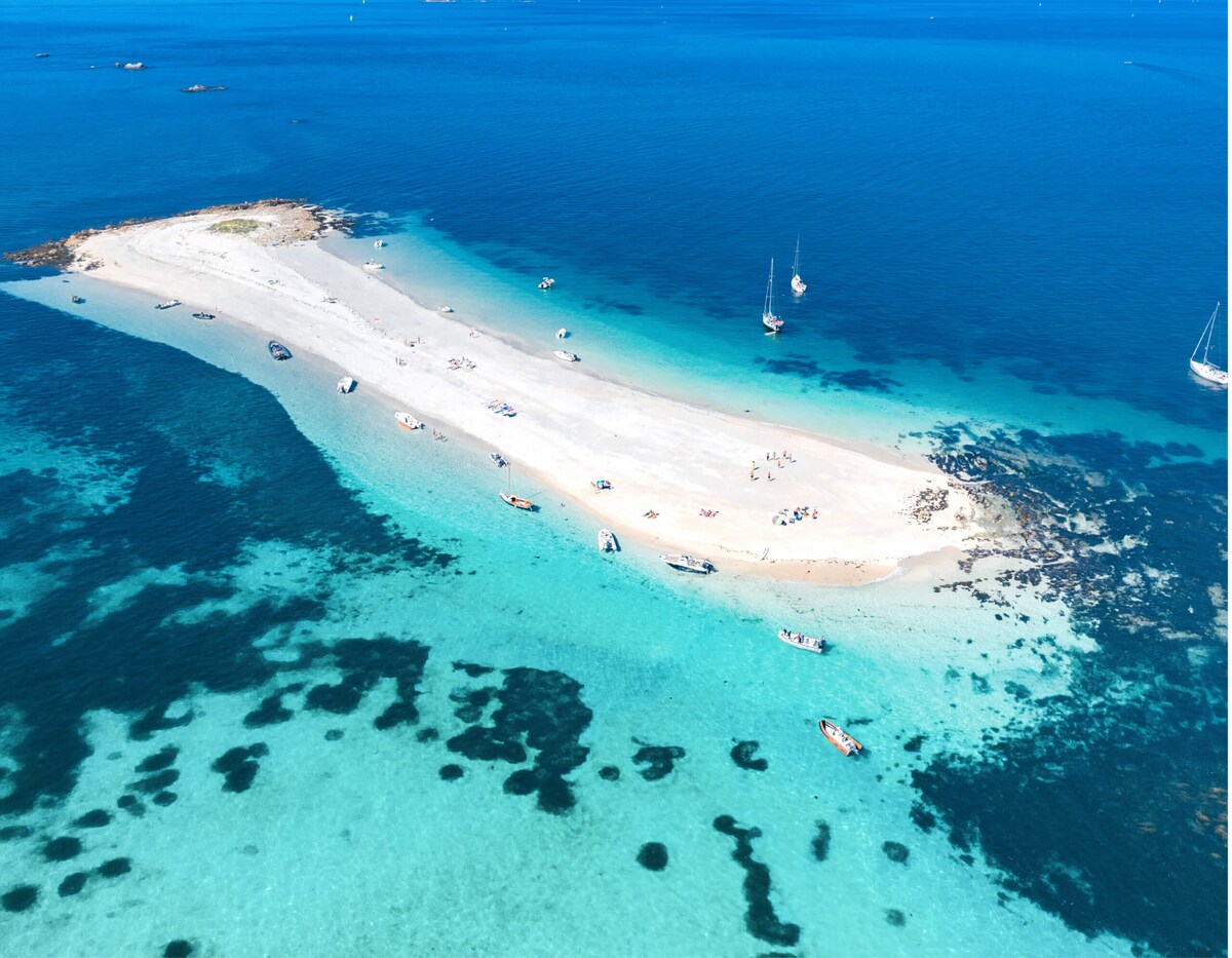 An aerial view captures a sandy islet surrounded by clear turquoise waters. Small boats are scattered around, and sunbathers can be seen on the beach. The gentle waves create a serene atmosphere, while the vibrant colors of the sea contrast with the white sand.