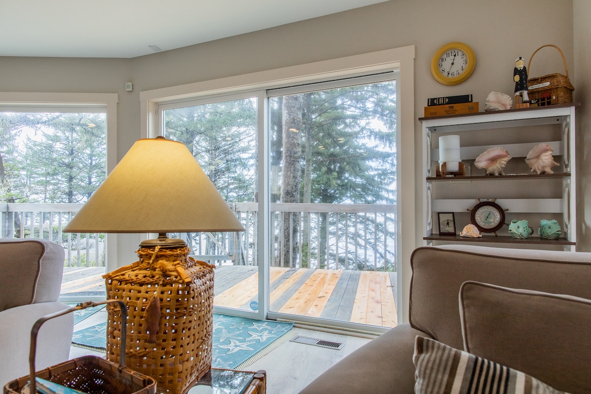 A cozy living area features a wicker lamp atop a patterned table. Light streams through expansive glass doors that open to a wooden deck overlooking trees and the ocean. A shelf displays seashells and decorative items, adding subtle coastal charm to the space.