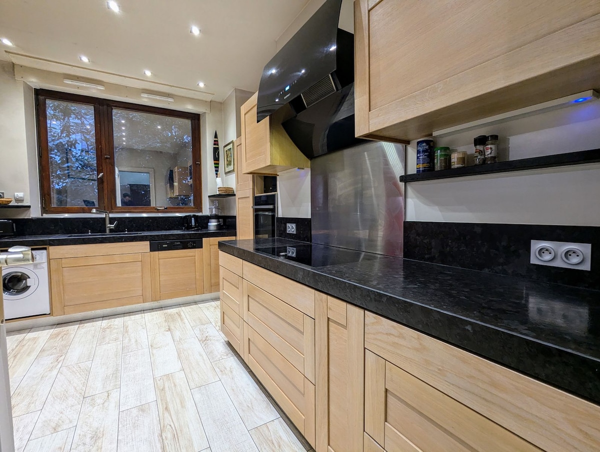 A modern kitchen with wooden cabinetry and sleek black countertops is featured. The space includes a large window that provides natural light, while stainless steel appliances are visible. Shelves display various cooking essentials, and the floor is covered with light-colored tiles.