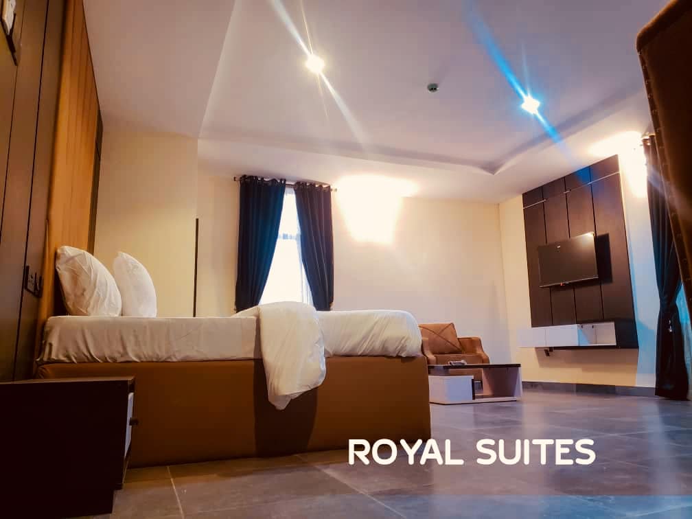 A spacious room is featured with a large bed dressed in white linens, complemented by dark curtains framing a window. A comfortable seating area includes a sofa, and a wall-mounted TV offers entertainment options. Soft lighting enhances the room's serene ambiance.