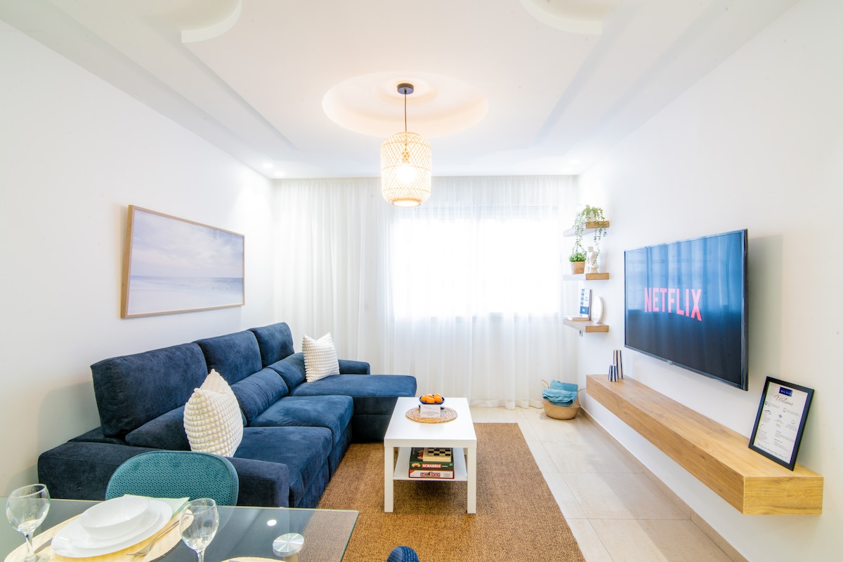 A bright living area is featured, showcasing a spacious dark blue sofa with decorative pillows. A light wood coffee table sits at the center, adorned with snacks. A wall-mounted TV displays the Netflix logo, while a large window allows natural light to flow in.