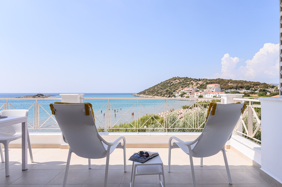 A serene terrace features two white lounge chairs overlooking a clear blue sea, with gentle waves and a distant sandy beach in view. A small coffee table holds a magazine, while a light breeze seems to create a relaxing atmosphere.