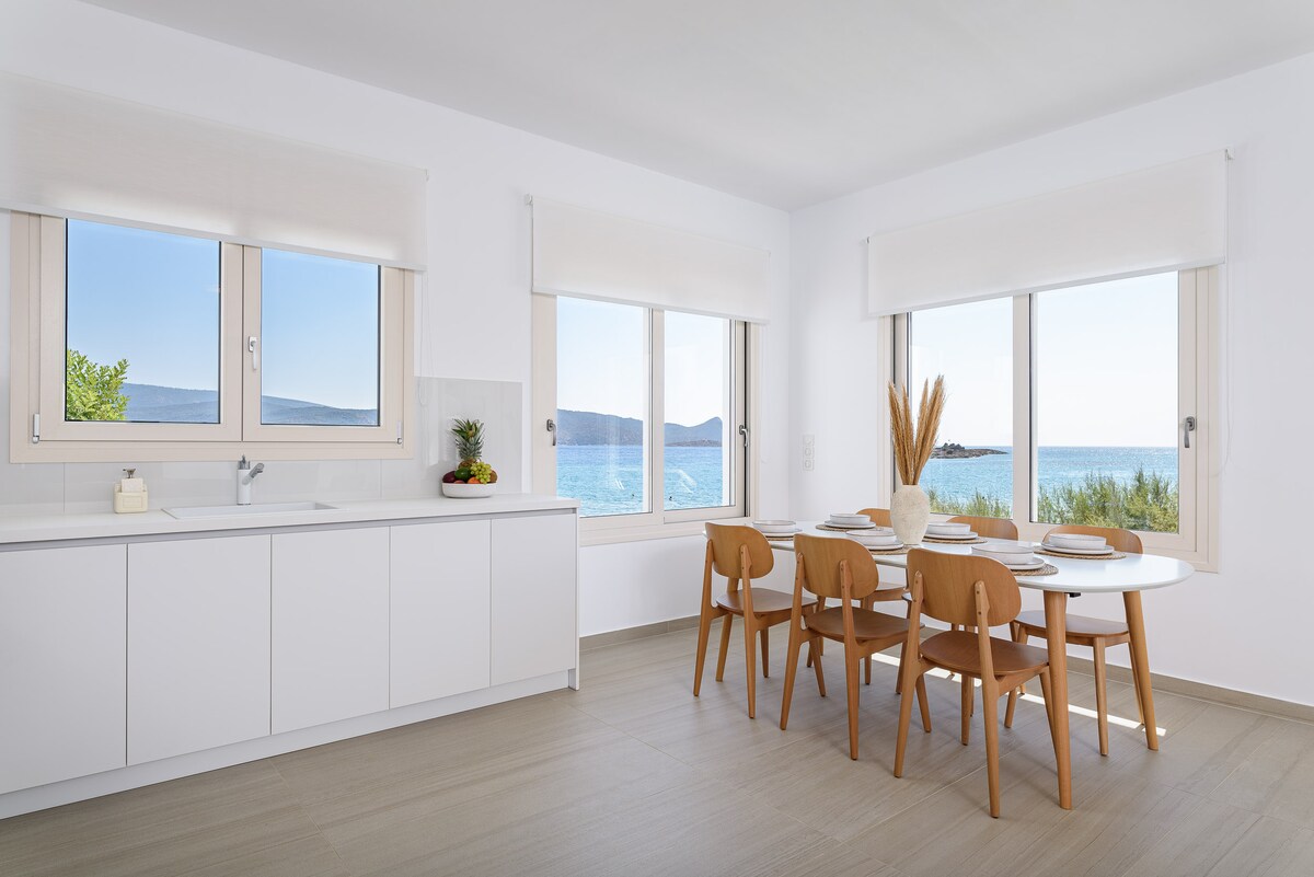 A bright dining area features a wooden table surrounded by matching chairs, with an expansive view of the sea visible through large windows. The space is complemented by minimalist cabinetry and soft natural light, enhancing the open and airy feel.