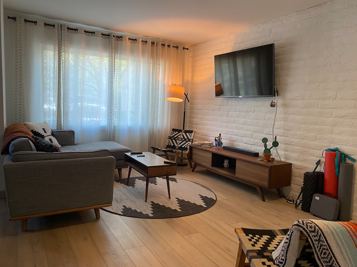 A modern living area features a comfortable sectional sofa and a wooden coffee table set on a patterned rug. Large windows are dressed with sheer curtains, allowing natural light to fill the space. A mounted flat-screen TV and stylish accent chair complete the room.