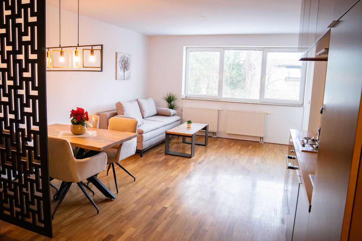 A bright living area is highlighted by a comfortable gray sofa and a wooden dining table surrounded by soft chairs. Natural light streams through a large window, illuminating the warm wooden floor. Greenery is present in the form of potted plants, enhancing the tranquil atmosphere.