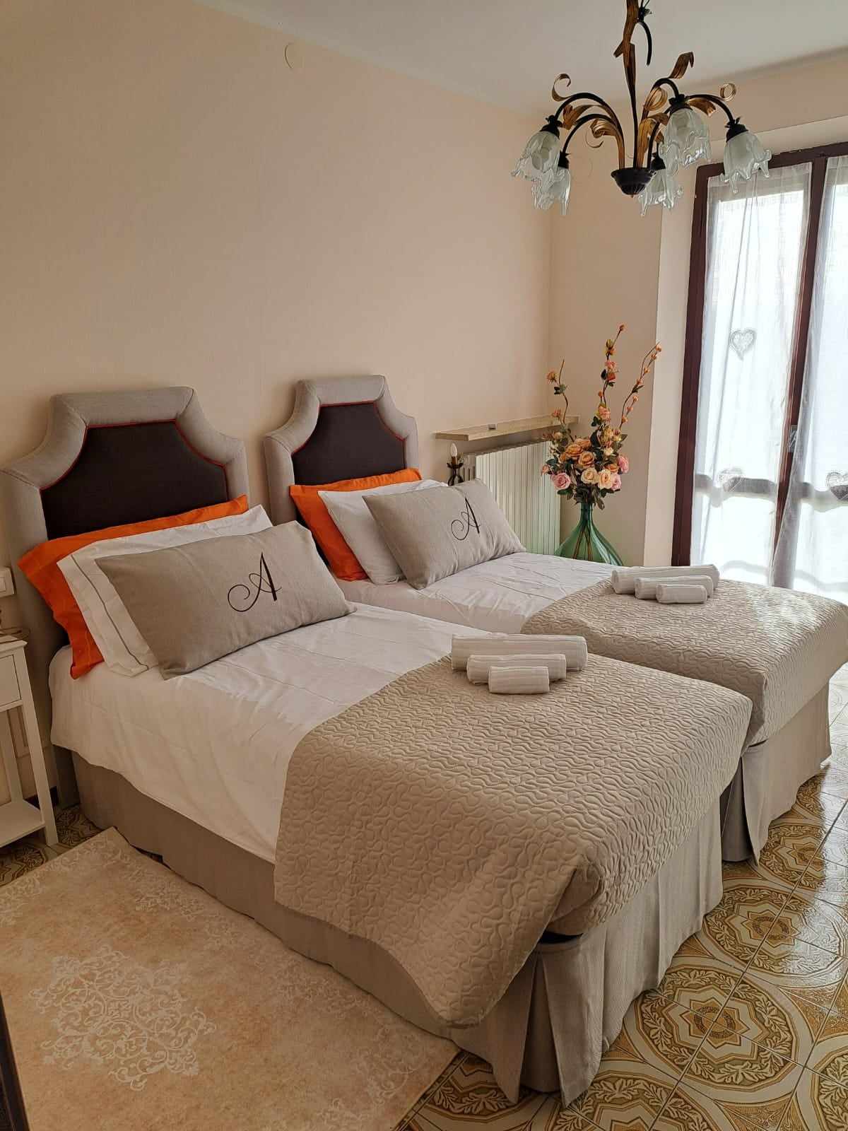 A comfortable guest room features two individual beds with elegant headboards, dressed in neutral bedding. Soft orange accents adorn the pillows, complemented by a decorative chandelier overhead. Natural light filters through double doors that open to a delightful view, enhancing the inviting ambiance.