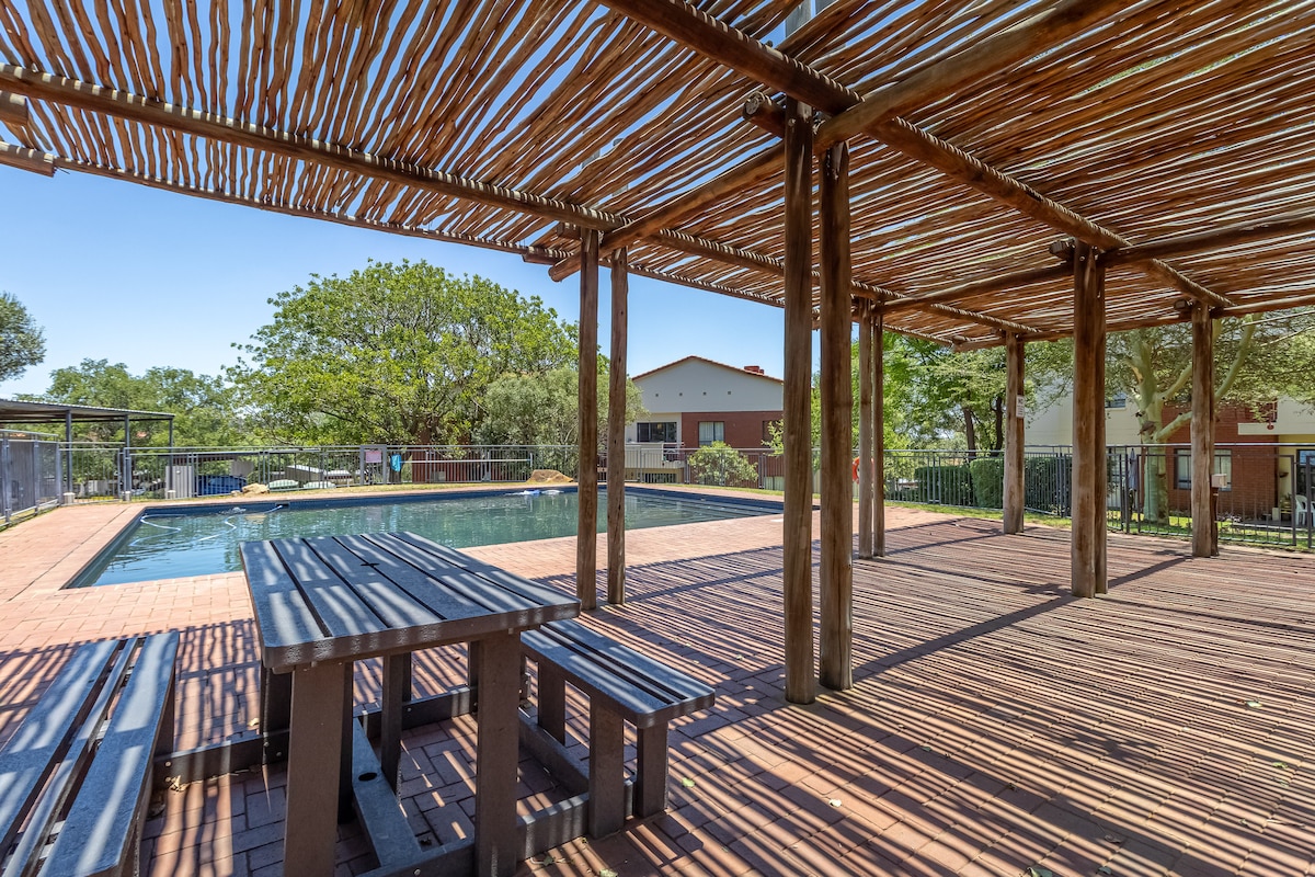 A shaded outdoor area is seen, featuring a wooden structure with a roof made of slats. A rectangular swimming pool is visible in the background, surrounded by a paved patio. A long wooden table with attached benches is situated in the foreground.