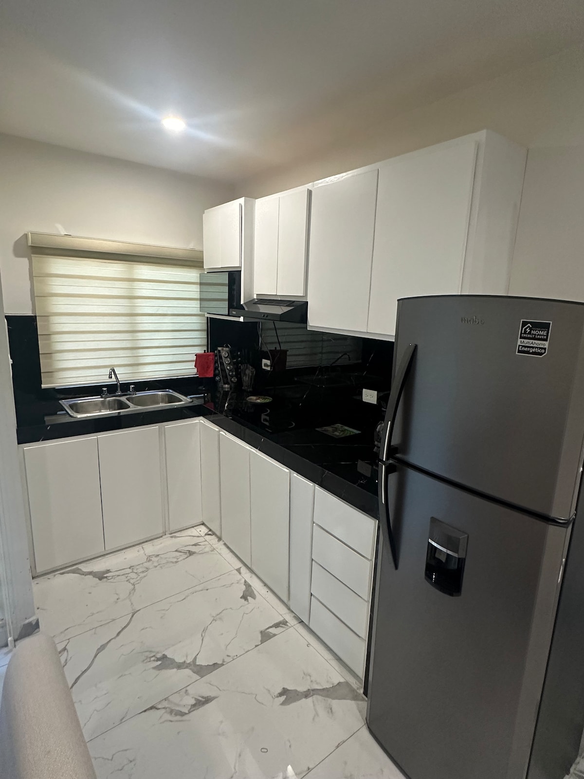 A modern kitchen is presented with sleek white cabinetry and a black countertop. Stainless steel appliances, including a refrigerator, are visible. A double sink is positioned under a window with light-filtering shades, and marbled flooring adds a touch of elegance to the space.