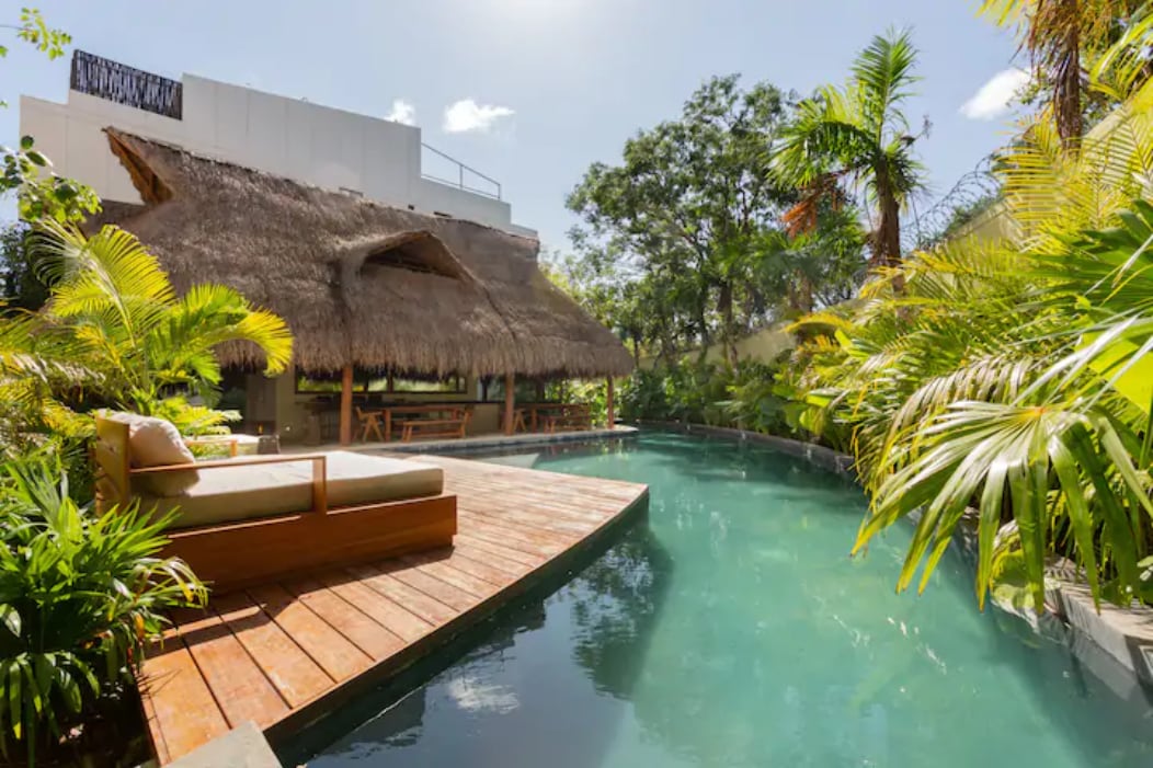 A serene outdoor space features a natural swimming pool surrounded by lush tropical vegetation. A wooden deck extends over the water, with a cozy sitting area beneath a thatched roof structure, offering a harmonious integration with nature.