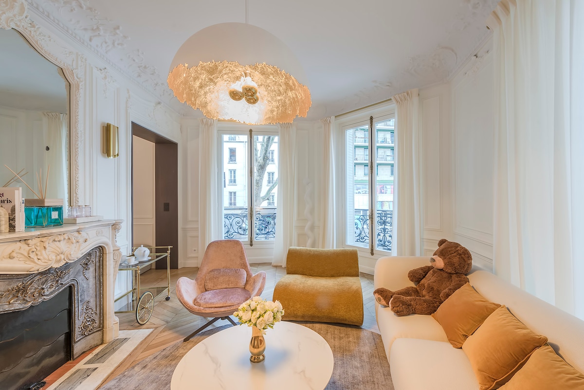 A sophisticated living area is seen, featuring a plush sofa complemented by decorative pillows. A round coffee table is central, accompanied by a teddy bear. Elegant curtains frame large windows, allowing natural light to illuminate the space, while an ornate chandelier adds a touch of luxury.
