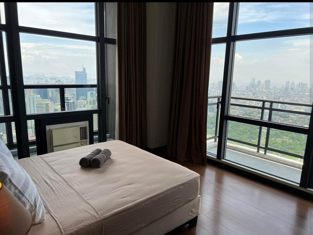 ركن غرفة نوم رئيسية مريح مع شرفتان تطلان على مدينة ماكاتي وخليج مانيلا وغروب الشمس. تشمل حمامًا ملحقًا وطاولة عمل وكرسي وخزانة ومكواة مع طاولة كي ومجفف شعر وسريرًا مرنًا بحجم كوين لراحة مطلقة.