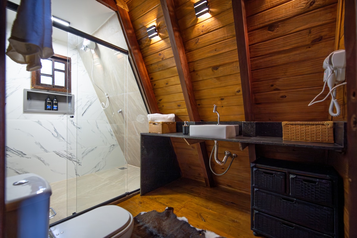The bathroom features wooden walls and a sloped ceiling, creating a rustic ambiance. A glass shower enclosure is complemented by modern fixtures. A sleek sink is positioned on a dark countertop, with woven baskets providing storage. Towels are neatly arranged, adding to the organized appearance.
