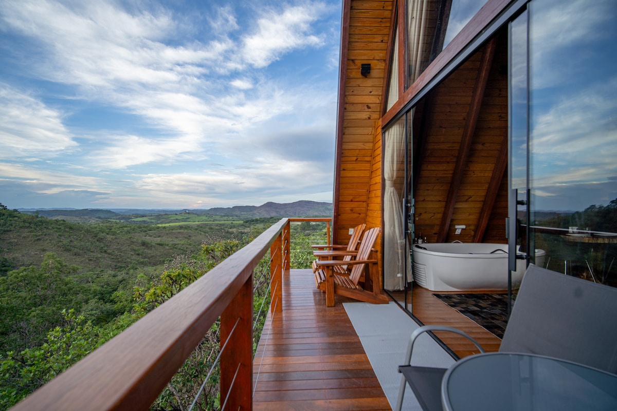 A wooden deck features a pair of adirondack chairs and a table, offering views of surrounding lush mountains and natural scenery. The chalet's large glass doors connect the outdoor space to the interior, allowing light to enter the cozy living area.
