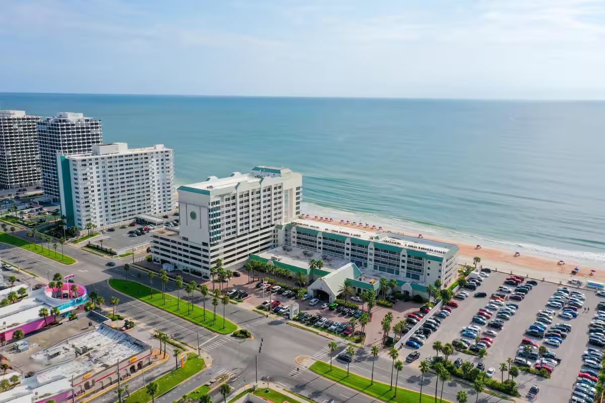 An aerial view captures the Oceanfront Hideaway and surrounding area, with a wide stretch of sandy beach meeting the calm ocean. Nearby, the prominent hotel structure is shown alongside a parking lot filled with vehicles, while lush greenery lines the streets.