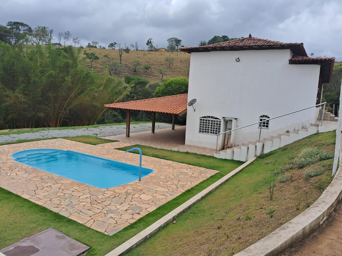 A well-maintained outdoor area features a swimming pool surrounded by a paved patio. The house is set against a backdrop of gently sloping hills, with a covered seating area nearby. Lush greenery is visible, enhancing the tranquil environment.