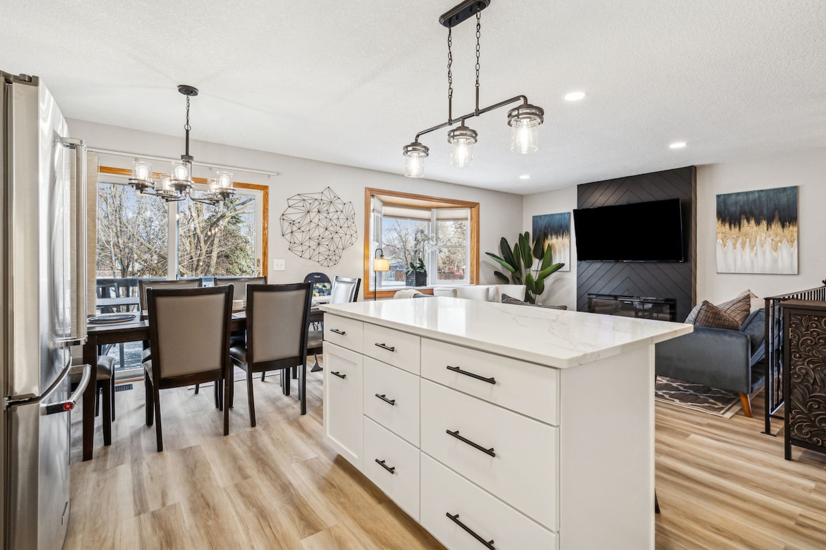 A bright and open kitchen features a large white island with contrasting dark hardware. A dining area is visible, with a wooden table surrounded by six upholstered chairs. Natural light floods the space through large windows, highlighting the modern decor and a wall-mounted television.
