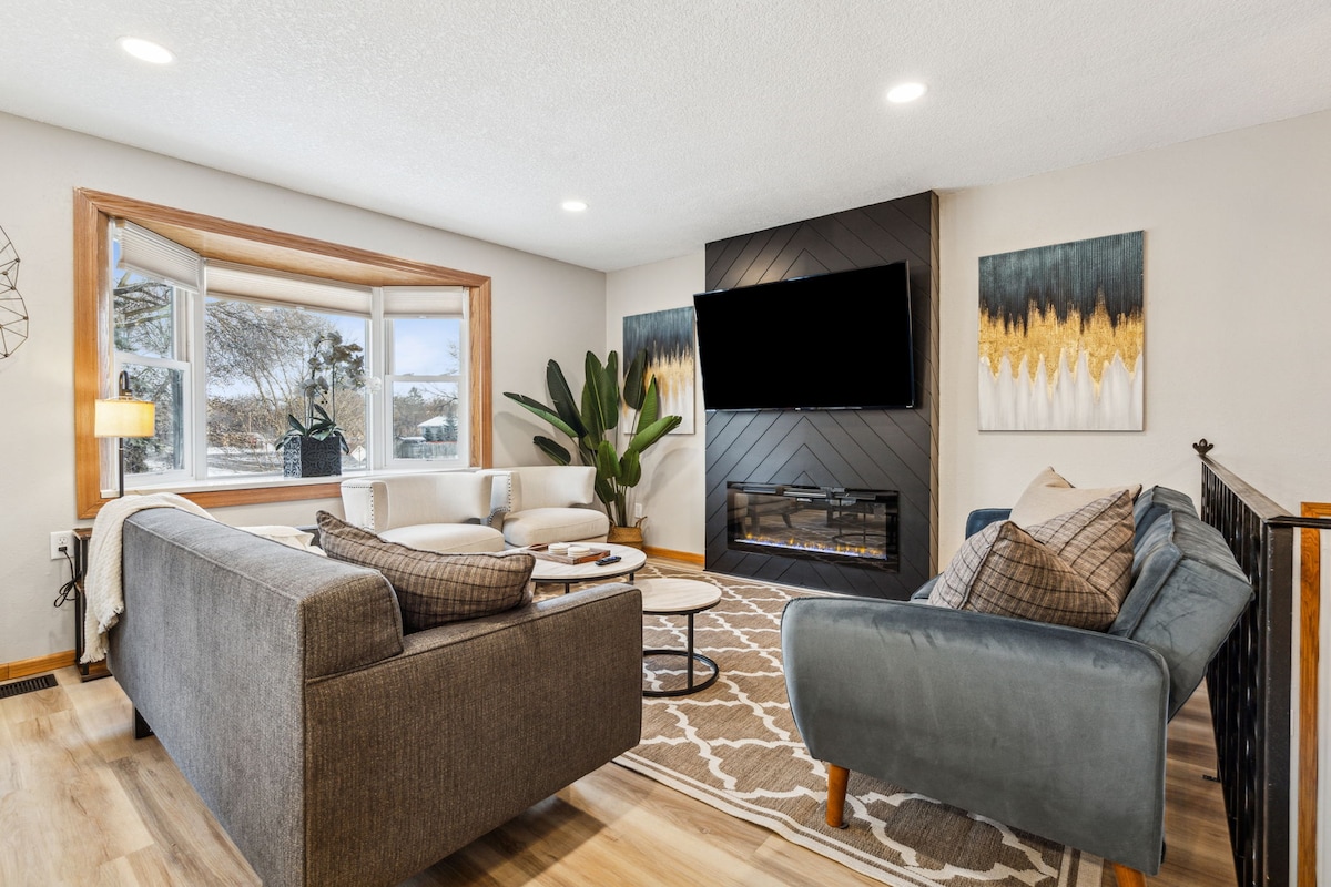 A cozy living area features a comfortable sofa and armchair arranged around a circular coffee table. A modern fireplace and large windows offer natural light, while abstract wall art adds a touch of style. A leafy plant provides a refreshing element in the space.