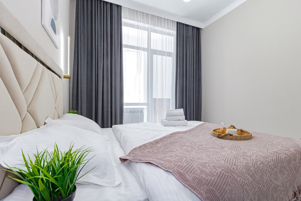 A well-appointed bedroom features a spacious bed layered with soft white linens and a light pink throw. A decorative tray holds pastries, placed alongside a small green plant. Natural light filters through sheer curtains, illuminating the neutral-toned walls and adding warmth to the environment.