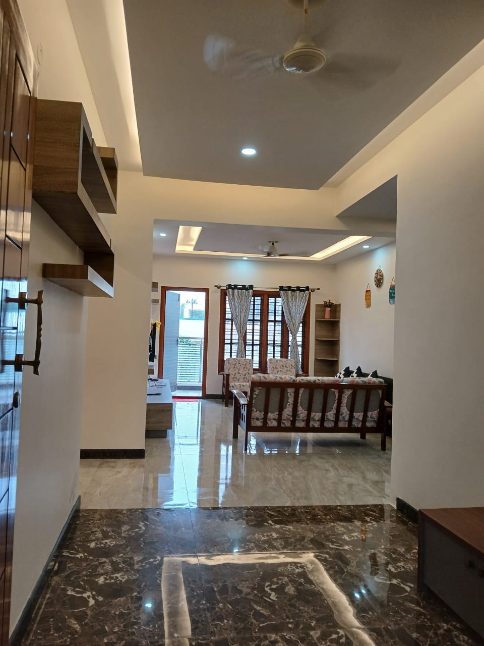 A spacious hallway showcases a polished marble floor leading to a bright living area. Natural light filters through closed wooden shutters, illuminating the light-colored walls. A wooden bookshelf and a sofa with patterned cushions are visible in the background, inviting relaxation.
