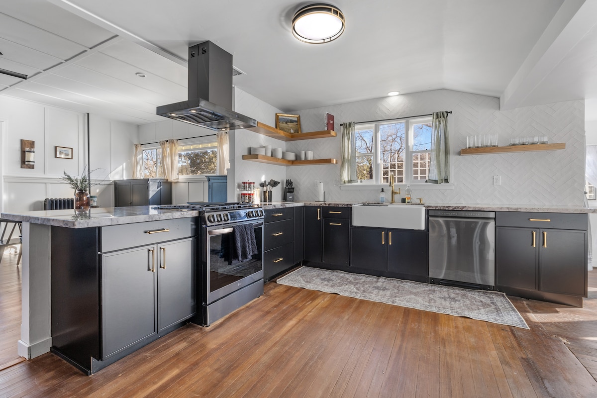 The kitchen features modern black cabinetry and a large farmhouse sink under a window. A stylish range and hood are included, along with a variety of countertop appliances. The hardwood floor adds warmth, while an area rug complements the space.