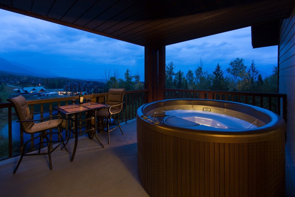 A private balcony features a circular hot tub and a small table with two chairs. The views include trees and distant mountain peaks under a twilight sky, creating a peaceful setting for relaxation.
