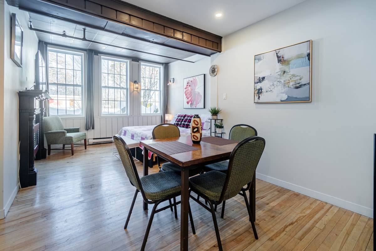 A bright and airy loft space features large windows that allow natural light to fill the room. A cozy seating area is complemented by a dining table with four chairs. Artwork decorates the wall, and a small decorative plant adds a touch of greenery.