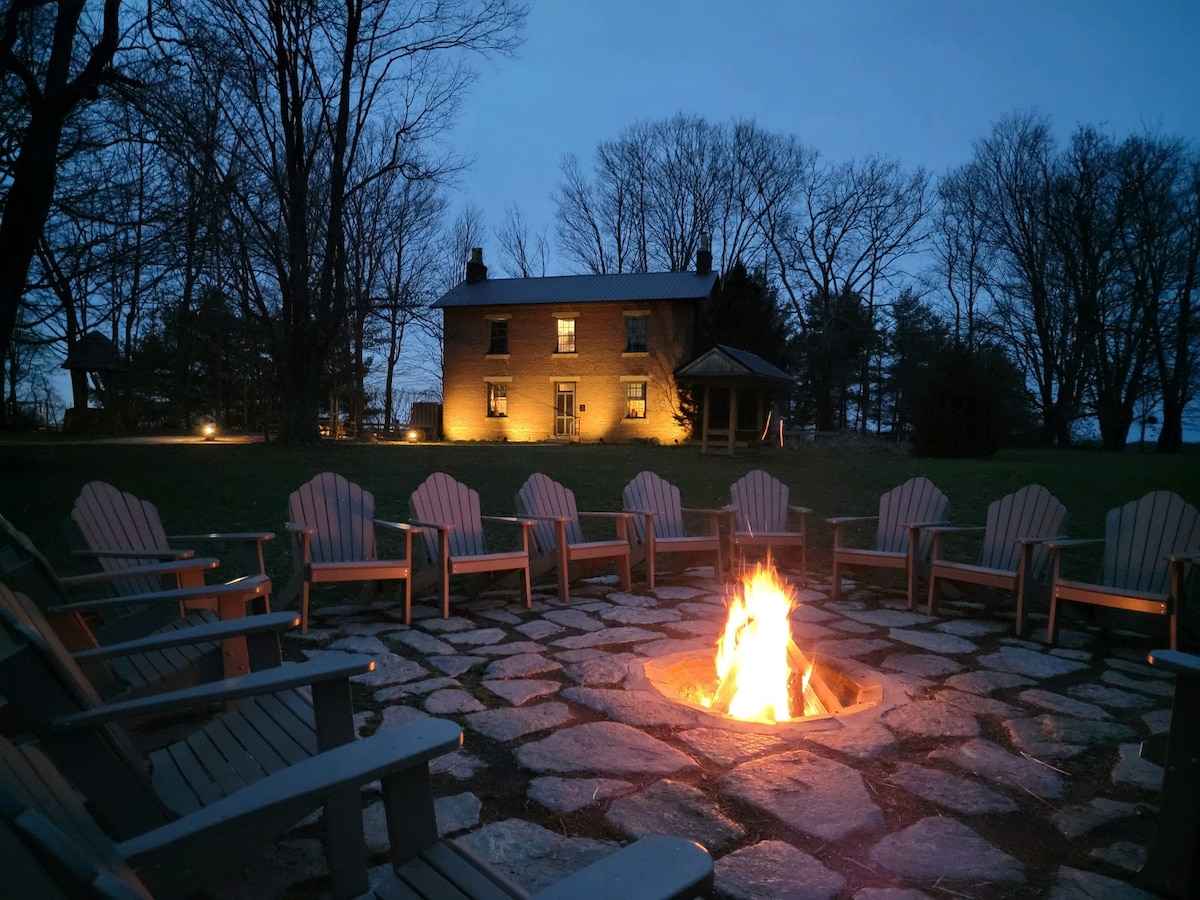 A fire pit surrounded by sturdy wooden chairs is illuminated at dusk. The warm glow of the flames contrasts with the cool evening air. In the background, a two-story brick building is softly lit, set against a backdrop of trees.