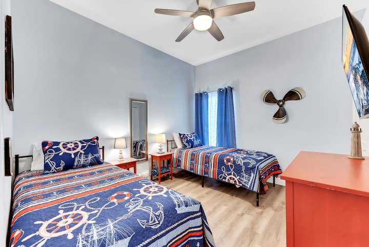 Esta habitación de temática náutica cuenta con dos camas individuales con ropa de cama marítima y detalles rojos. El televisor Roku de 43pulgadas, el ventilador de techo y la ventana garantizan la comodidad en este acogedor refugio costero.
