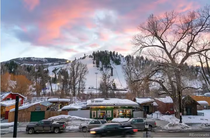 A serene winter landscape is presented, with soft, pastel-colored clouds overhead. Snow-covered hills and tree-lined slopes are visible in the distance. Below, a cozy scene features charming local buildings positioned along a road, with vehicles subtly moving through the snowy setting.
