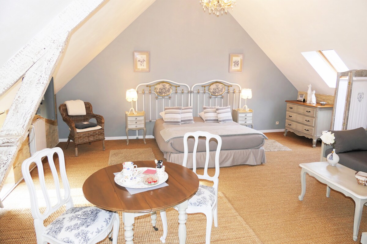 A spacious room features sloped ceilings and soft blue walls. A large double bed is positioned centrally with striped linens. A rustic wicker chair is situated beside the bed. A round wooden table with two chairs holds a breakfast set. Warm light is provided by bedside lamps.
