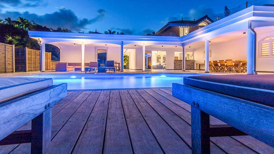 An inviting outdoor space features a spacious wooden deck, with plush lounge chairs positioned near a serene pool. Soft blue lighting enhances the area, while modern architecture showcases a seamless flow between indoor and outdoor living.