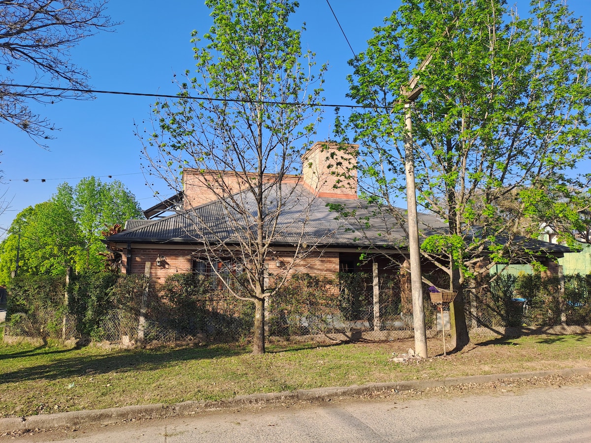 The exterior view shows a single-story home surrounded by green trees and a well-maintained yard. A fence encloses the property, highlighting the brick structure with a sloped roof. The clear blue sky complements the natural surroundings.