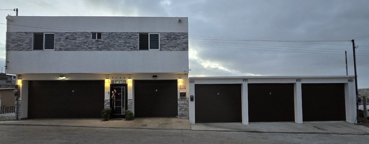 A modern building façade features a two-story structure with stone accents and multiple windows. Attached garages are visible, providing secure parking options. The entrance is well-lit, enhancing safety and accessibility. A landscaped area with potted plants is located near the entrance.