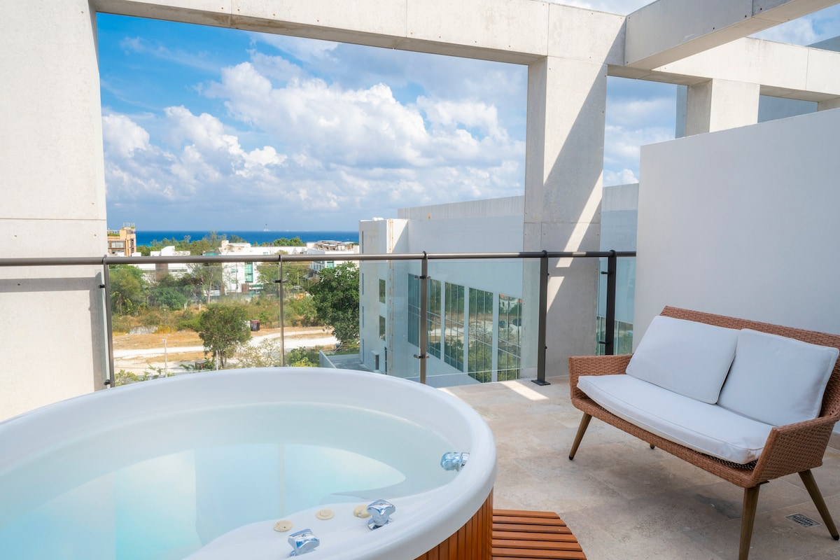 A private terrace features a hot tub with a view of the ocean and surrounding landscape. A modern outdoor seating area with a plush white sofa is included, providing a relaxing space to enjoy the scenery. The area is bright and airy, framed by minimalist architecture.