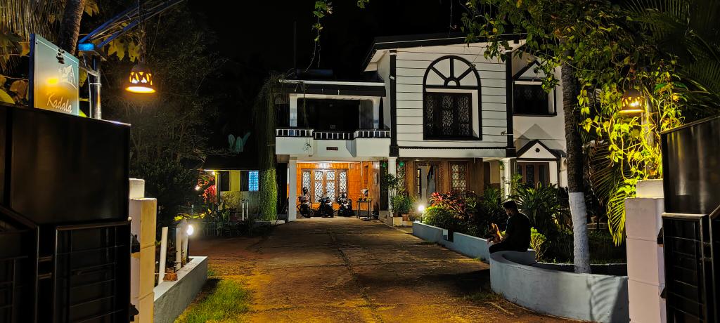 An inviting view of the entrance to the home is displayed at night, with warm lighting illuminating the pathway. Lush greenery and colorful plants line the sides of the driveway, leading to a welcoming front porch with decorative features.