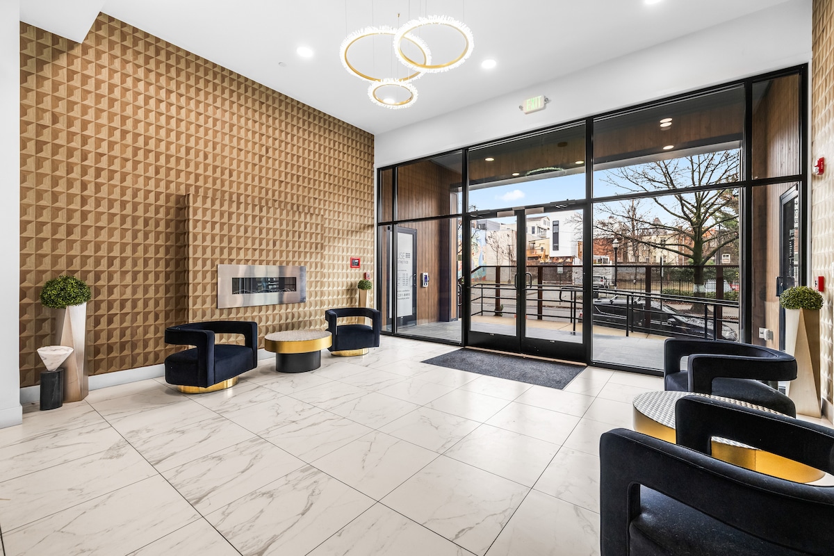 A modern lobby welcomes guests with large glass doors providing natural light. A geometric wall design enhances the space, complemented by a contemporary seating arrangement featuring dark chairs and circular tables. Potted plants add a touch of greenery, creating an inviting entryway.