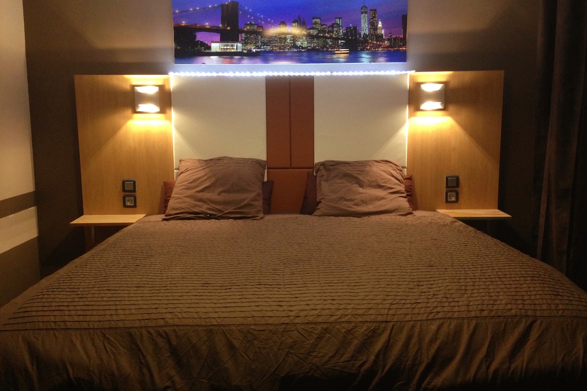 A spacious bed is featured with textured bedding and two pillows positioned neatly on each side. Soft lighting fixtures are mounted on the headboard, illuminating the area. A city skyline artwork is displayed above the bed, adding a modern touch to the room.