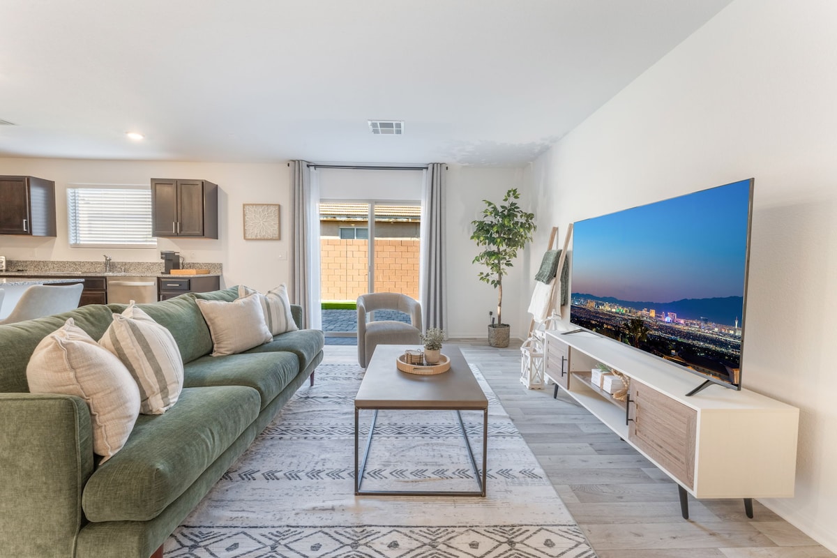 A spacious living area features a green sectional sofa and a coffee table in front of a contemporary TV stand. Soft light filters through large windows, illuminating the neutral tones of the space. A potted plant adds a touch of greenery, and a decorative area rug defines the seating area.