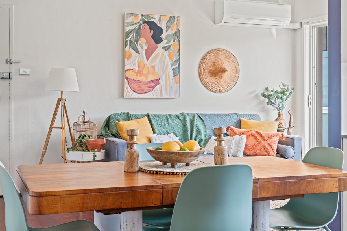 A cozy living area features a colorful artwork above a grey sofa adorned with various cushions. A wooden dining table is set with a bowl of fruit and surrounded by green chairs. Natural light filters in through a nearby window and an air conditioning unit is visible.