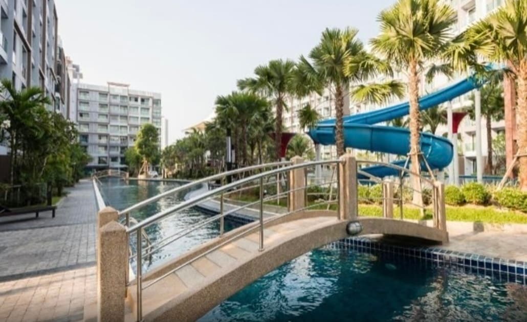 A smooth stone bridge spans across a calm pool, surrounded by palm trees and greenery. A blue water slide can be seen in the background, enhancing the outdoor recreational atmosphere of the area. Spacious pathways lead alongside the water for accessible enjoyment.
